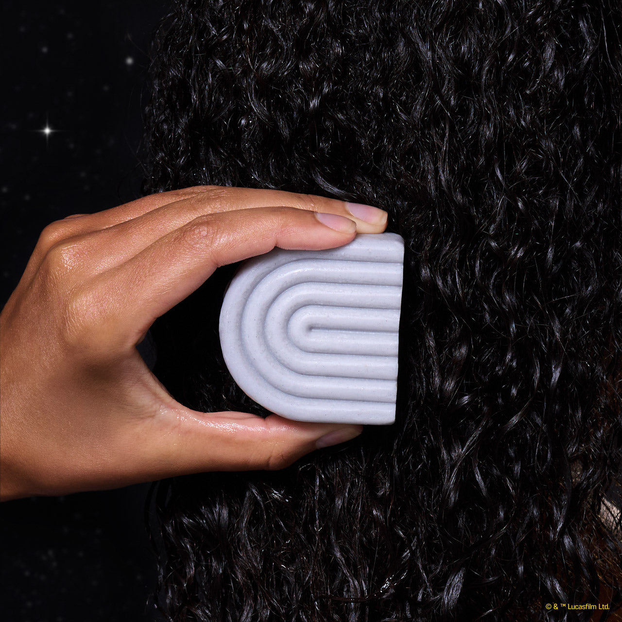 Hand holding KITSCH x Star Wars™ silver-grey S-shaped Rice Water Protein Solid Conditioner Bar (R2-D2 edition) against dark curly hair — in-use lifestyle shot with galaxy starfield backdrop — Strengthening formula For thin, fine, & damaged hair