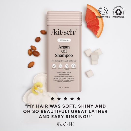 5-star customer review flat-lay for KITSCH Repairing Argan Oil Shampoo on white background styled with argan nuts, grapefruit slice, and white flower — Katie W.: "MY HAIR WAS SOFT, SHINY AND OH SO BEAUTIFUL! GREAT LATHER AND EASY RINSING!!" — Cruelty-Free, Post-Consumer Packaging