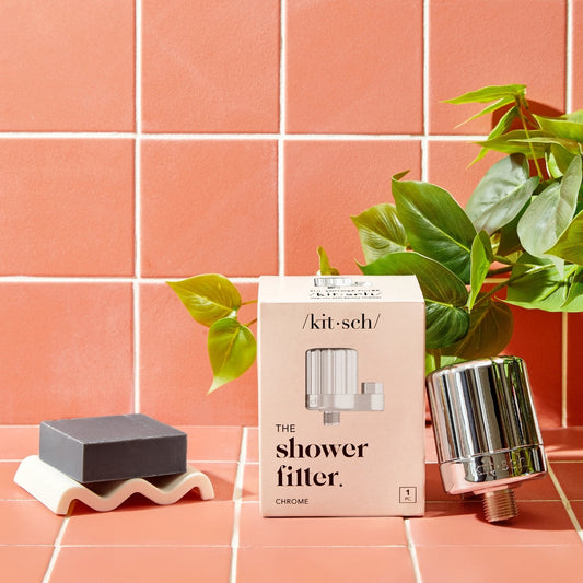 Kitsch Shower Filter in Chrome with product packaging displayed on a bathroom counter beside a soap dish and plant.