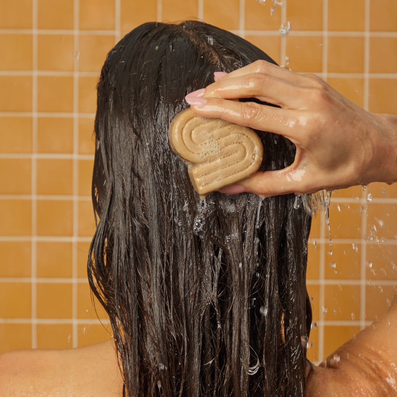 A Model with long wet dark hair washes their hair with a beige, wavy-S shaped shampoo bar against a tiled wall background.