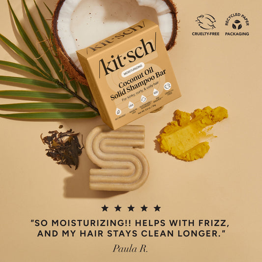 Kitsch coconut oil shampoo bar with palm leaf and coconuts on a beige background. There is a Leaping Bunny Cruelty-free badge and a Recycled paper packaging badge on the top right corner. On the bottom of the image, there is a customer review that says "so moisturizing! helps with frizz and my hair stays clean longer"