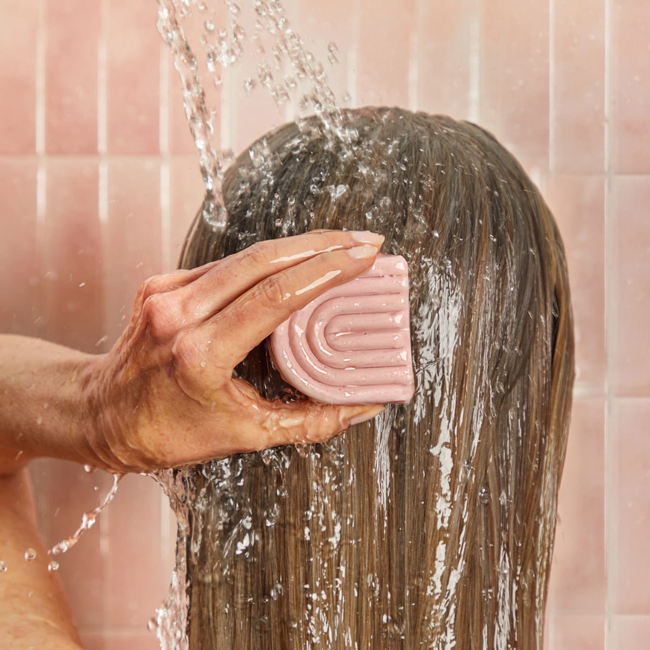 A close-up of a model washing their hair with water running over it, holding a pink Conditioner bar against the scalp.