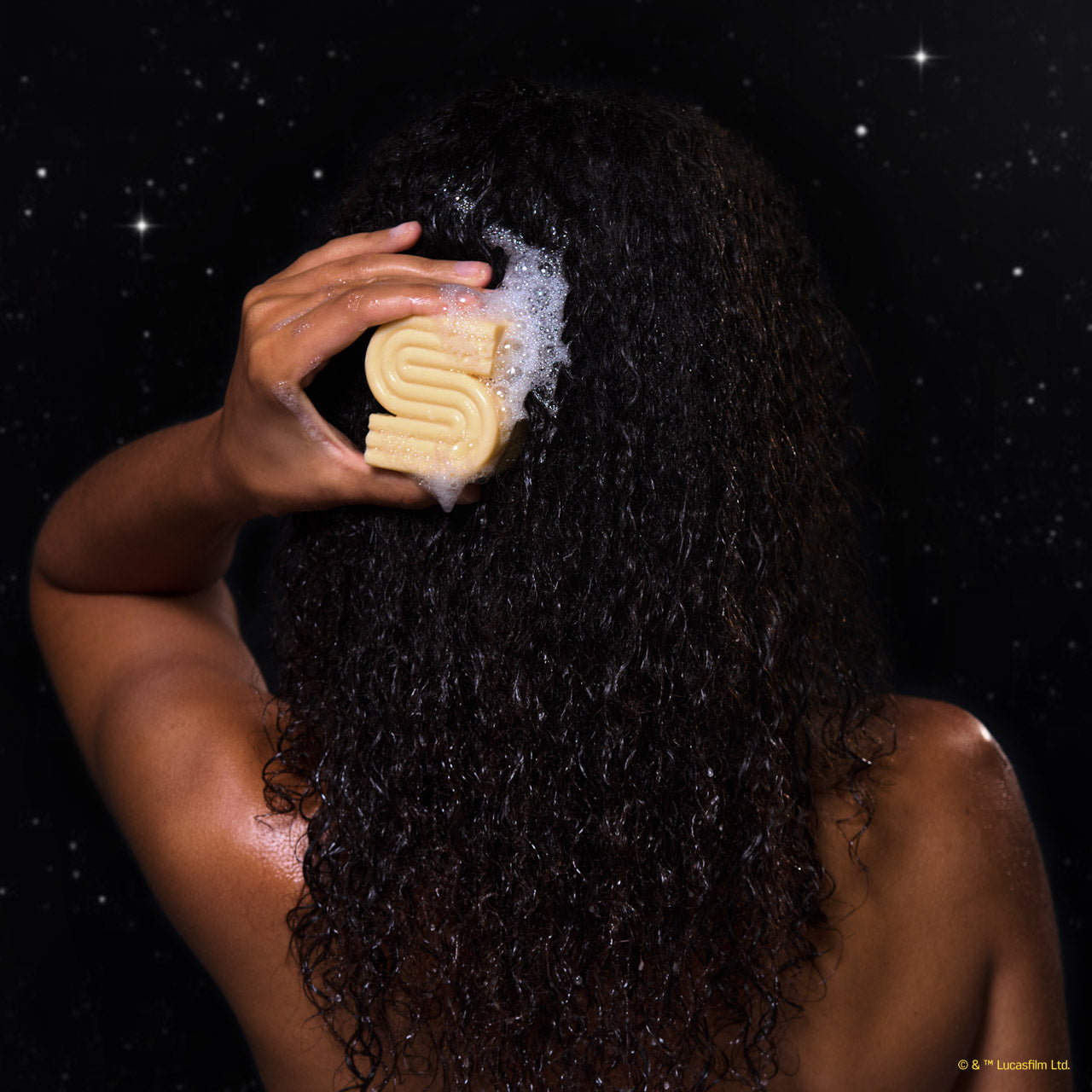 Back view of model applying lathered KITSCH x Star Wars™ honey-gold S-shaped Rice Water Protein Solid Shampoo Bar (C-3PO edition) to dark curly wet hair — lifestyle in-use shot against galaxy starfield backdrop — Strengthening formula For thin, fine, & damaged hair
