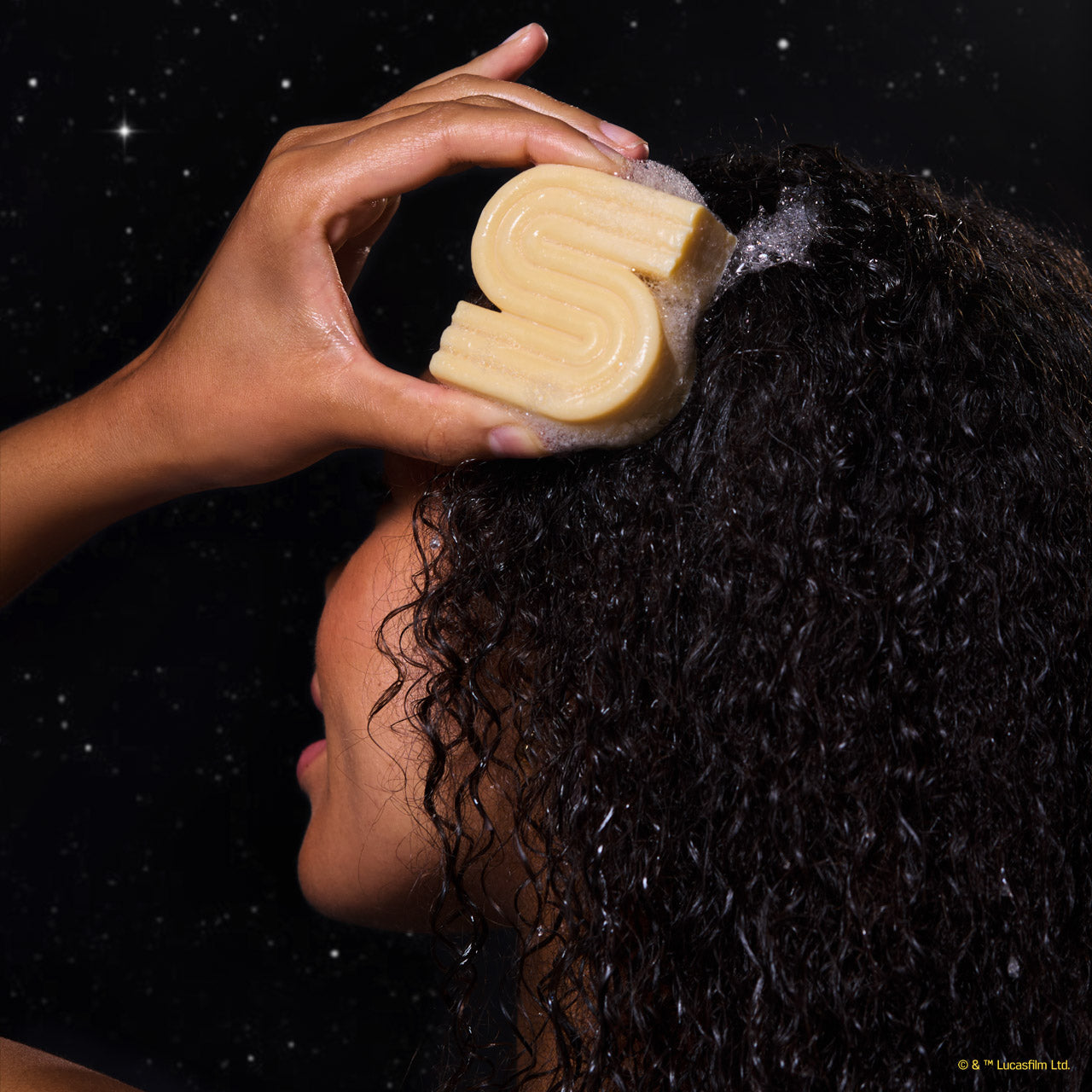 Model applying KITSCH x Star Wars™ honey-gold S-shaped Rice Water Protein Solid Shampoo Bar (C-3PO edition) directly to dark curly hair — in-use lifestyle model shot against galaxy starfield backdrop — Strengthening formula For thin, fine, & damaged hair