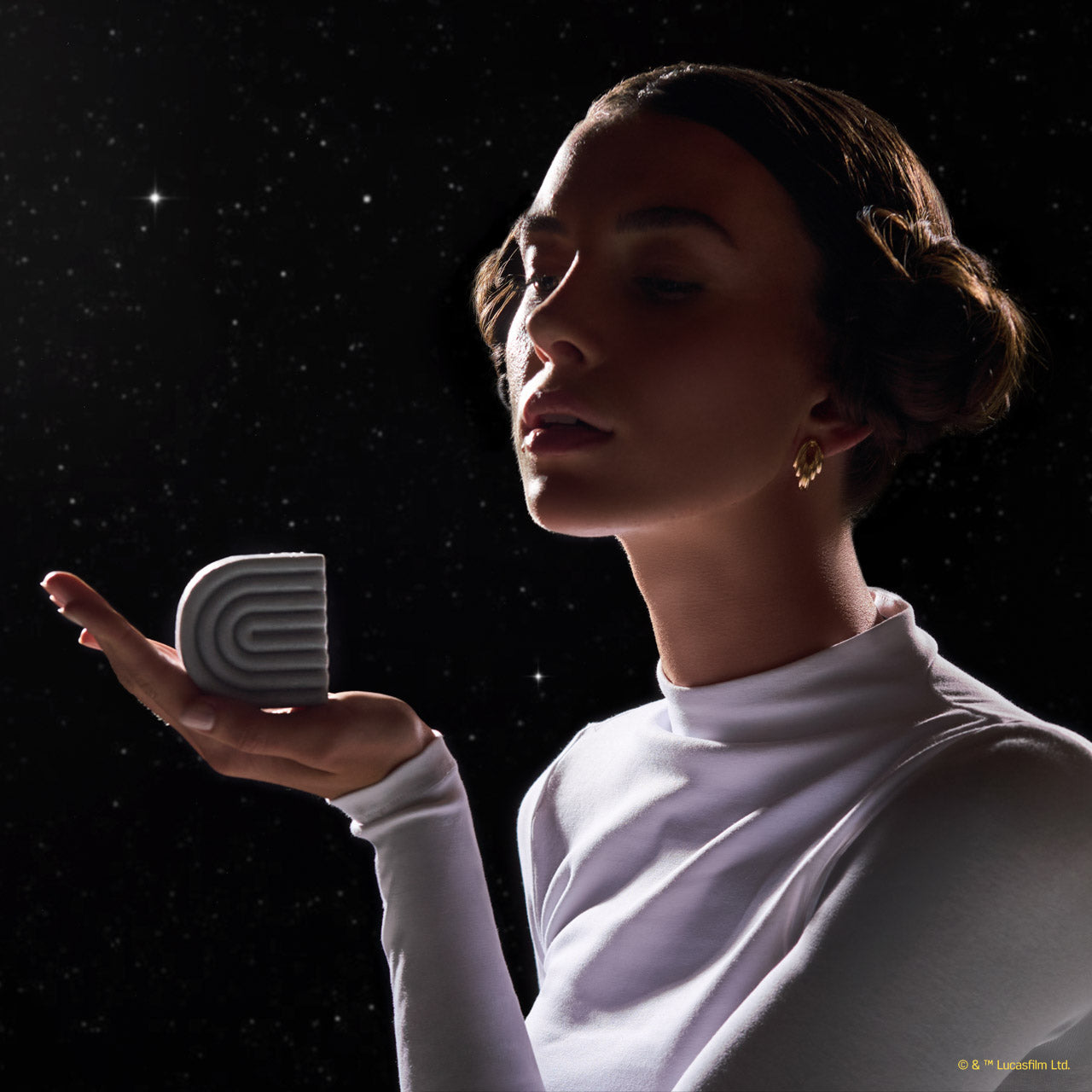 Female model in white turtleneck holding KITSCH x Star Wars™ silver-grey S-shaped Rice Water Protein Solid Conditioner Bar (R2-D2 edition) in outstretched palm — elegant lifestyle product shot against galaxy starfield backdrop — Strengthening formula For thin, fine, & damaged hair