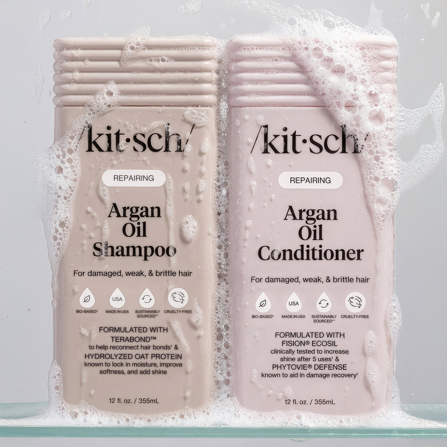 KITSCH Repairing Argan Oil Shampoo 12 fl. oz. and Conditioner 12 fl. oz. bottles in dynamic water and foam splash — both labeled REPAIRING, For damaged, weak, & brittle hair — Shampoo formulated with Terabond™ & Hydrolyzed Oat Protein; Conditioner formulated with Fision® EcoSil & PhytoVie® Defense