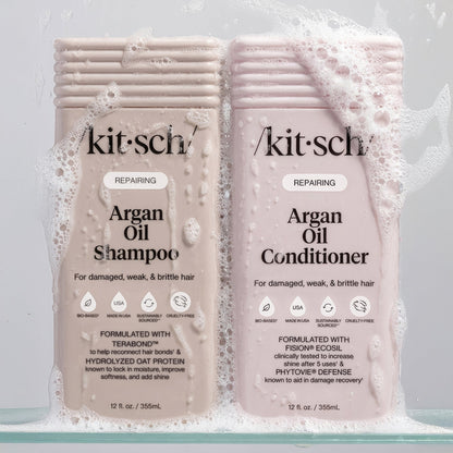 KITSCH Repairing Argan Oil Shampoo 12 fl. oz. and Conditioner 12 fl. oz. bottles in dynamic water and foam splash — both labeled REPAIRING, For damaged, weak, & brittle hair — Shampoo formulated with Terabond™ & Hydrolyzed Oat Protein; Conditioner formulated with Fision® EcoSil & PhytoVie® Defense