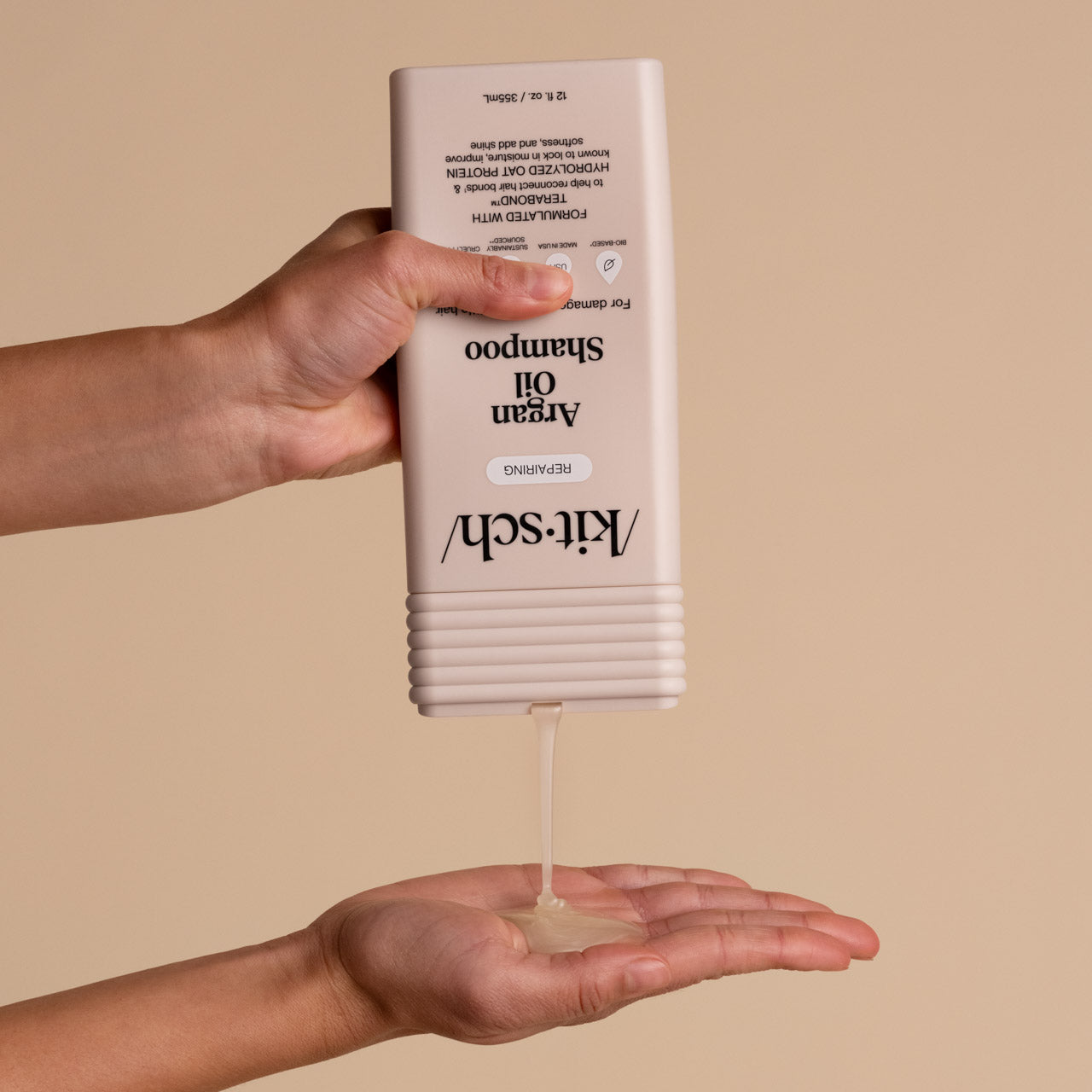 Hands dispensing KITSCH Repairing Argan Oil Shampoo — bottle held upside down pouring shampoo into open palm — product application lifestyle shot for Repairing formula for damaged, weak, & brittle hair with Terabond™ & Hydrolyzed Oat Protein