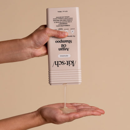 Hands dispensing KITSCH Repairing Argan Oil Shampoo — bottle held upside down pouring shampoo into open palm — product application lifestyle shot for Repairing formula for damaged, weak, & brittle hair with Terabond™ & Hydrolyzed Oat Protein