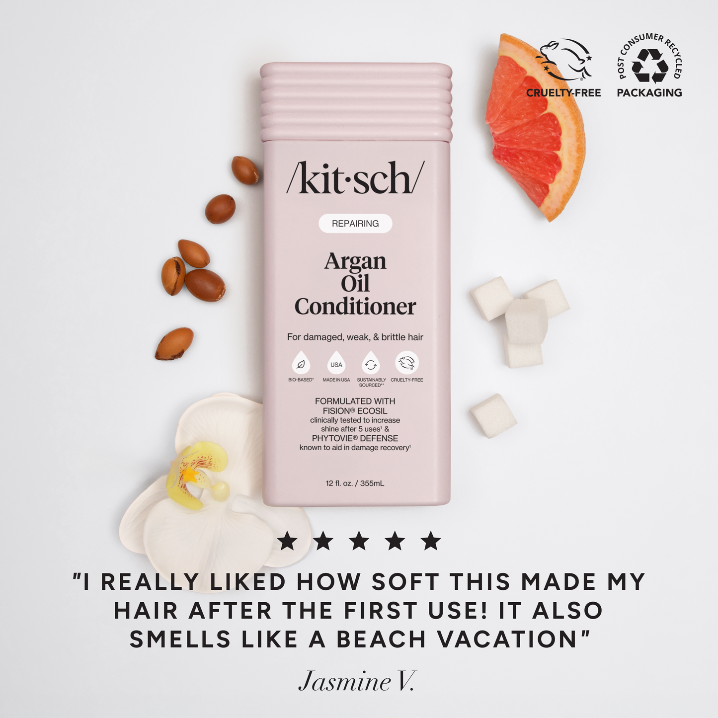 5-star customer review flat-lay for KITSCH Repairing Argan Oil Conditioner on white background styled with argan nuts, grapefruit slice, and white flower — Jasmine V.: "I REALLY LIKED HOW SOFT THIS MADE MY HAIR AFTER THE FIRST USE! IT ALSO SMELLS LIKE A BEACH VACATION" — Cruelty-Free, Post-Consumer Packaging