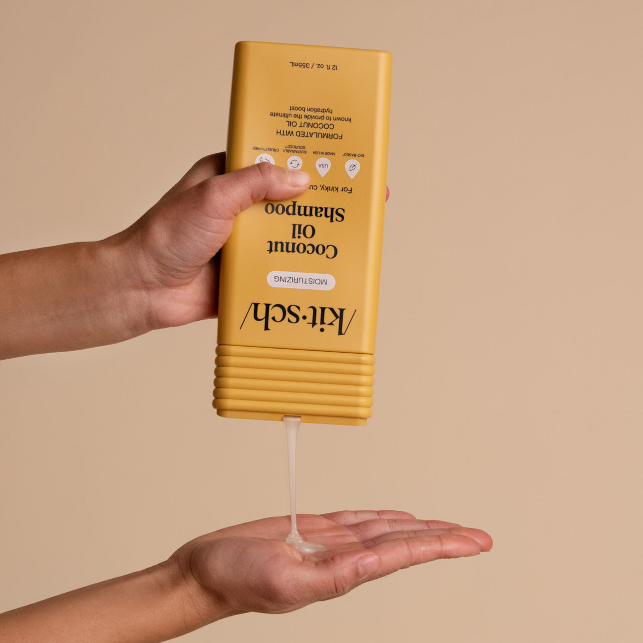 Hand dispensing KITSCH Moisturizing Coconut Oil Shampoo from yellow bottle into palm, beige background