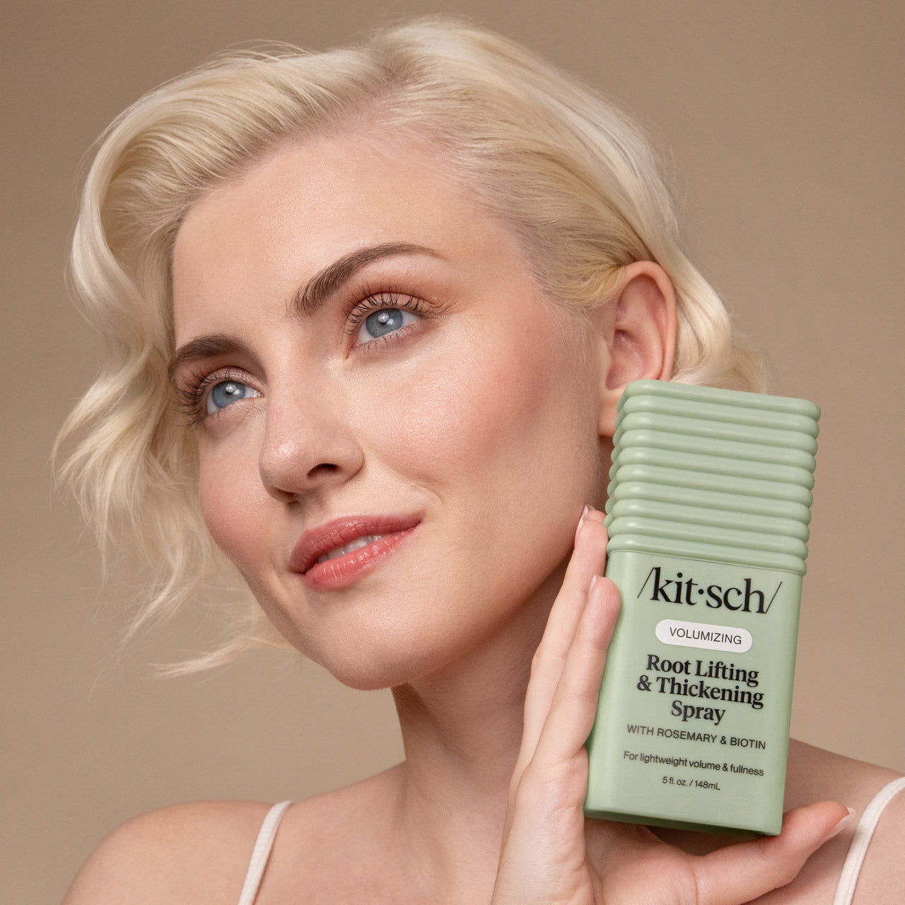 KITSCH Root Lifting & Thickening Spray — lifestyle portrait of female model with short blonde voluminous wavy hair, holding bottle beside cheek, warm beige background — For lightweight volume & fullness at your roots — 90% agreed did not weigh hair down or have a greasy feel — Infused with rosemary & Biotin to help promote the appearance of healthy hair