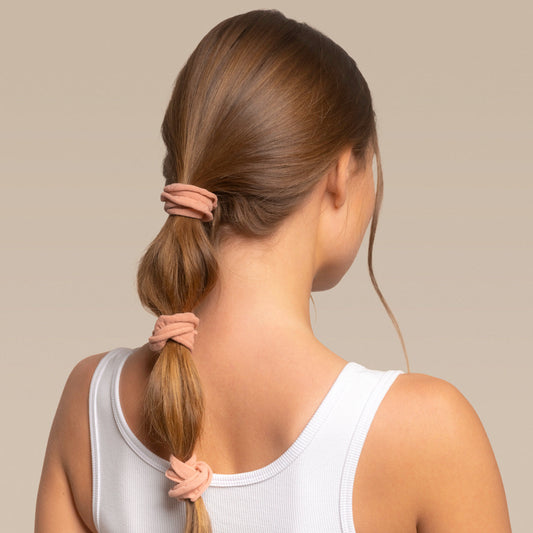 Woman with a bubble ponytail with 3 kitsch petal hair elastics wearing a white tank top against a beige background