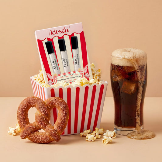 KITSCH Movie Theater Hair Perfume Trio Set — creative product shot on warm beige background — trio packaging set inside popcorn bucket surrounded by spilled popcorn, cinnamon pretzel and tall fizzy cola drink — Buttered Popcorn, Fizzy Cola & Cinnamon Pretzel scents — Smell like a snack with three delectable scents — Patented technology eliminates odors at the source — Sheer, versatile & easy to use