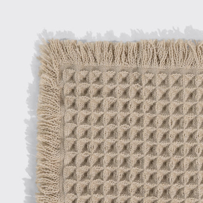 Close-up of a camel waffle-knit bath mat's corner, showing the woven texture and fringed trim.