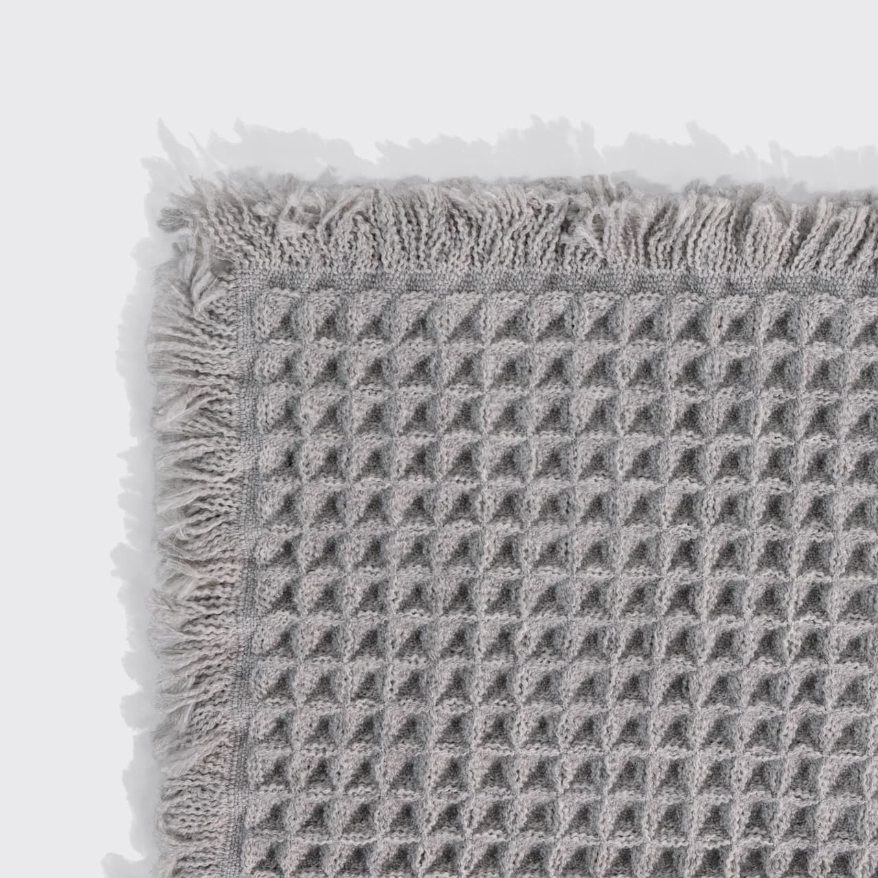 Close-up of a gray waffle-knit bath mat's corner, showing the woven texture and fringed trim.