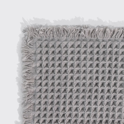 Close-up of a gray waffle-knit bath mat's corner, showing the woven texture and fringed trim.
