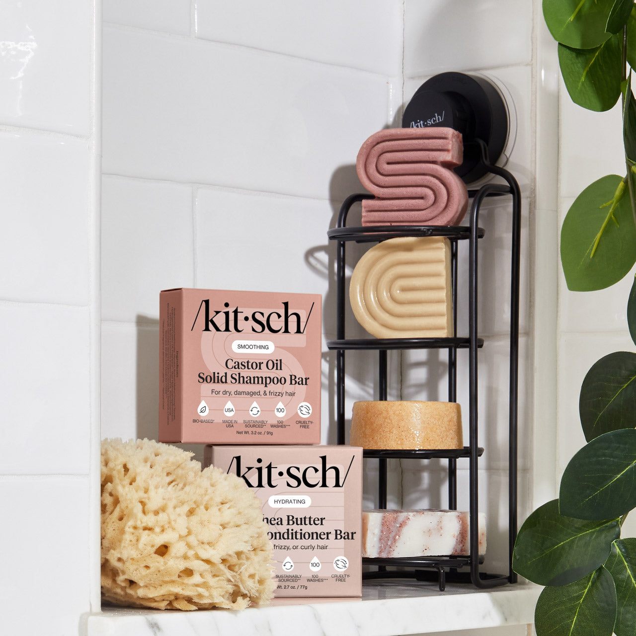 KITSCH Shea Butter Hydrating Solid Conditioner Bar — lifestyle bathroom shot with bar and box on black metal shower caddy — styled with KITSCH Castor Oil Solid Shampoo Bar, natural sea sponge, and tropical plant — white tile background — Richly hydrates dry, thirsty strands — PH balanced and safe for color-treated hair — Reduces single-use plastic