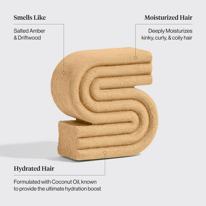 Beige textured shampoo bar with text describing its properties on a light gray background. Properties include: Smells like salted amber & driftwood, moisturized hair - deeply moisturizes kinky, curly, & coily hair, hydrated hair - formulated with coconut oil, known to provide the ultimate hydration boost