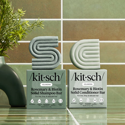 KITSCH Volumizing Rosemary & Biotin Solid Shampoo Bar and Conditioner Bar displayed alongside their packaging boxes on a green tile surface with rosemary plant and modern ceramic props — both bars labeled VOLUMIZING, For fine, limp, & delicate hair — bottle-free 2-bar combo set